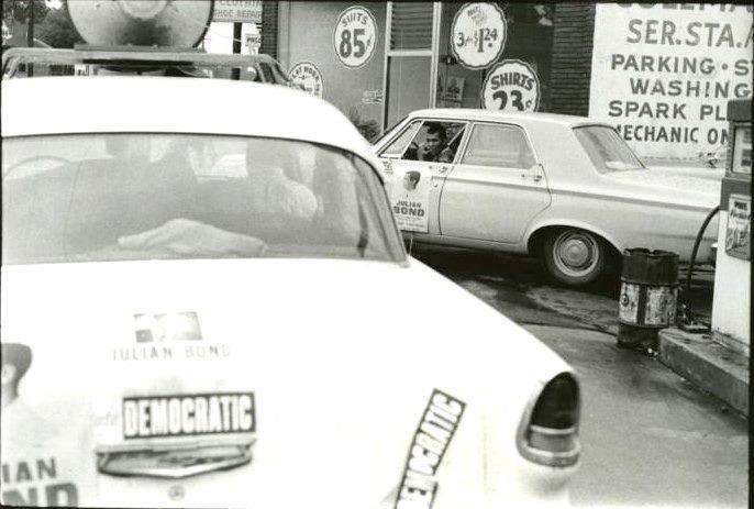 Two cars with Democratic bumper stickers sit in a parking lot, Ivanhoe Donaldson is in the driver's seat of the car in the background looking out the window.